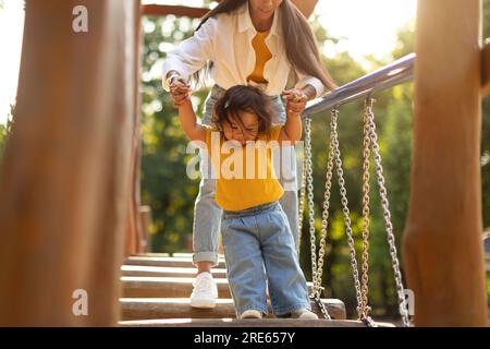 Korean Mommy Holding Daughter's Hands Walking Swinging Bridge On Playground Stock Photo