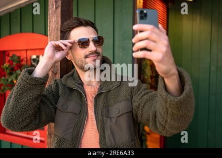 Handsome young man taking self portrait while walking through the city. Urban millenial with beard wearing sunglasses using smartphone and smiling carefree outdoors. Lifestyle concept. Stock Photo