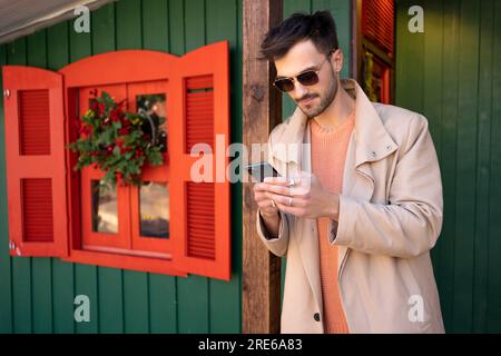 Stylish man wearing sunglasses and trench coat standing outside on Christmas market in European city. Using his smartphone to send holiday greetings enjoying festive atmosphere Stock Photo