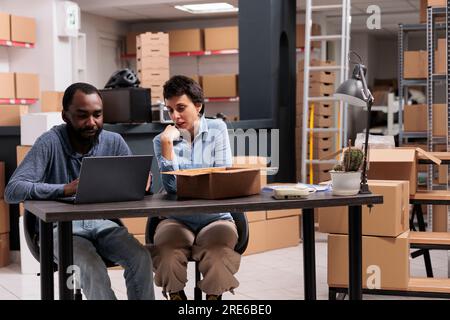Employees looking at cargo stock on laptop after finishing packing trendy clothes in carton boxes before delivering to clients working in warehouse. Concept of heavy industry manufacturing factory. Stock Photo