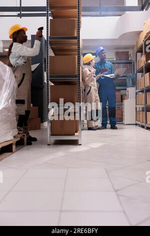 Factory warehouse diverse assistants team wearing protective overalls working. Shipping company storehouse colleagues managing parcels distribution and scanning containers bar codes Stock Photo
