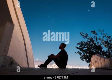 a man sits and looks at the night sky on the steps of the Ossuary Monument (Spomenik Kosturnica) in Veles, North Macedonia Stock Photo