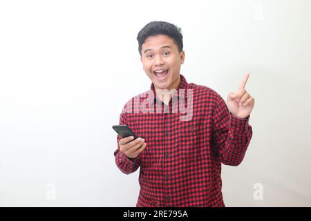 Portrait of attractive Asian man in casual plaid shirt standing against gray background, pointing away with his finger while using mobile phone. Adver Stock Photo