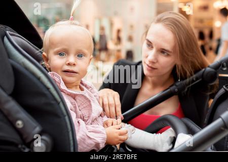 Close-up portrait of a small child in a carriage, blonde baby girl in pink clothes. Mom with a baby in a stroller walks around the mall, sat down next Stock Photo