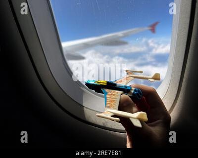 child hand holding toy plane in front of window of aircraft during flight. Cloudy sky and wing outside the airplane. Image with selective focus Stock Photo