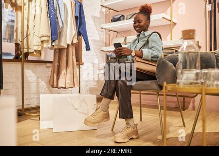 Full length portrait of black young woman using smartphone in clothing boutique and using mobile app, copy space Stock Photo