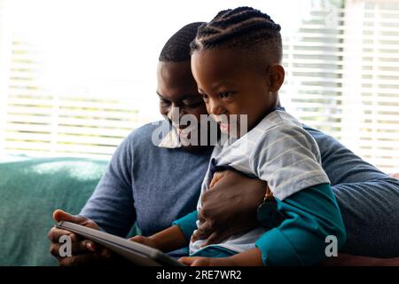 Happy african american father and son sitting in living room embracing and using tablet Stock Photo