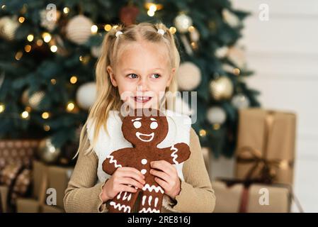 Merry Christmas and Happy Holidays. Child girl eating big iced ingerBread man cookies under Christmas tree Waiting for Christmas. Celebration New Year Stock Photo