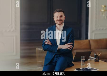 Satisfied bearded businessman in formal attire, stands confidently in office, arms folded, smiling pleasantly, content with work results. Stock Photo
