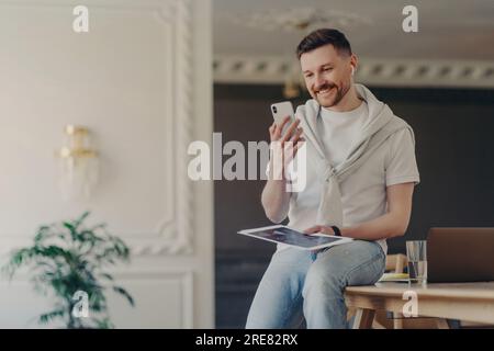 Bearded freelancer in coworking space, video calling on smartphone with wireless earphones, free high-speed internet, having an online conference with Stock Photo
