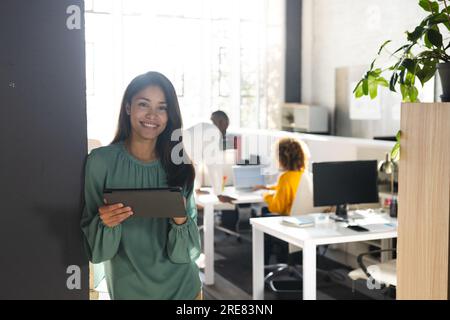 Portrait of happy biracial casual businesswoman using tablet over colleagues working in office Stock Photo