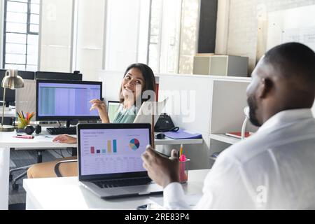 Happy diverse colleagues talking and using laptops with diagrams on screens in office Stock Photo