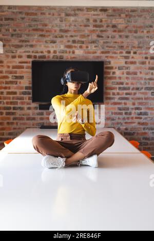 African american casual businesswoman sitting on table and using vr headset in office Stock Photo