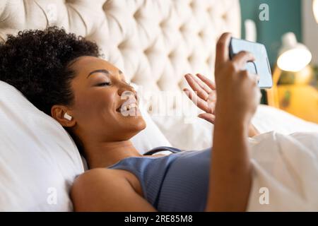 Happy biracial woman with earphones lying on bed and using smartphone in bedroom Stock Photo