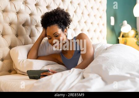Happy biracial woman with earphones lying on bed and using smartphone in bedroom Stock Photo