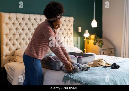Biracial woman packing suitcase and talking on smartphone in bedroom Stock Photo