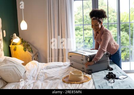 Happy biracial woman packing suitcase in bedroom Stock Photo