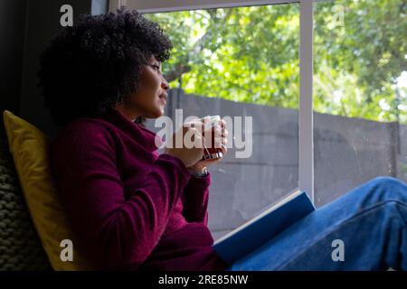 Thoughtful biracial woman sitting by window and drinking tea at home Stock Photo