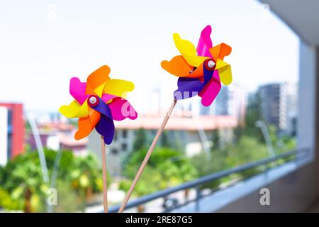 Colorful pinwheels spinning in the wind on a modern balcony with a blurry city background. Vibrant toy windmills capturing summer breeze. Stock Photo