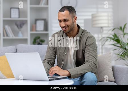 African american smiling young male student studying at home online remotely, sitting on sofa and typing on laptop. Stock Photo