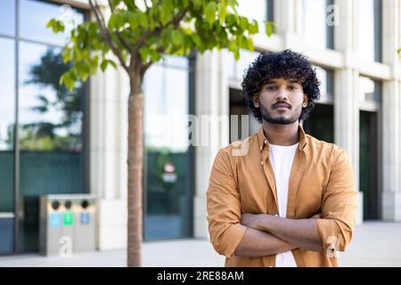 Portrait of a young male student, freelance designer, architect, engineer who stands with his arms crossed on the benches near the office center and looks seriously into the camera. Stock Photo