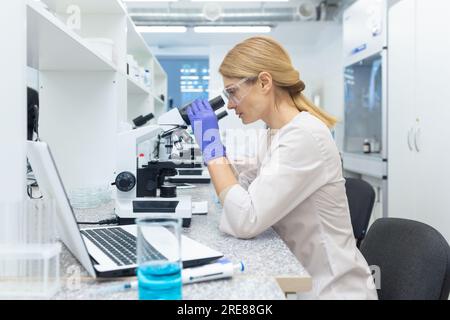 A young woman scientist, chemist, biologist sits in a laboratory at a table and examines substances under a microscope. Stock Photo