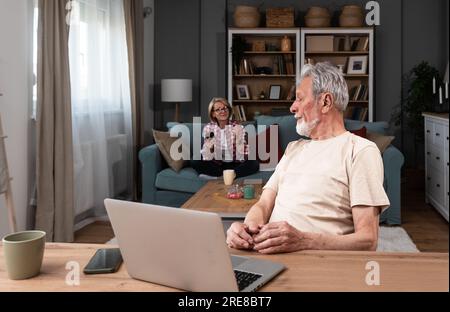 Senior couple argue at home while older retired military man trying to have video call on his laptop computer with army friend and his wife talking to Stock Photo