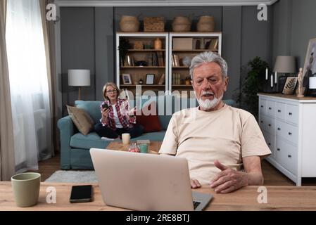 Senior couple argue at home while older retired military man trying to have video call on his laptop computer with army friend and his wife talking to Stock Photo