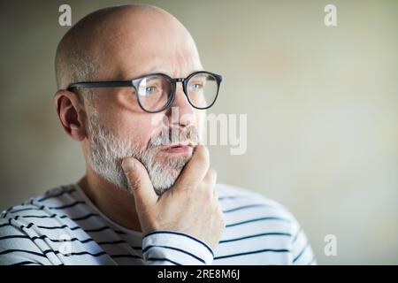 Serious pensive bald senior man with gray beard deep in thoughts touching chin and looking away Stock Photo