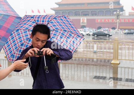 Chinese tourist photographer protecting himself from rain with umbrella while taking a photograph with digital camera on a wet day in Tiananmen Square, Beijing, China. Portrait of Chairman Mao Tse Tung watches from the walls of the Forbidden City. (125) Stock Photo