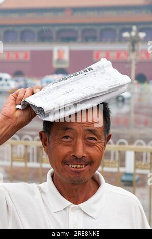 Chinese tourist protecting himself from rain with folded newspaper on a wet day in Tiananmen Square, Beijing, China. Portrait of Chairman Mao Tse Tung watches from the walls of the Forbidden City. (125) Stock Photo