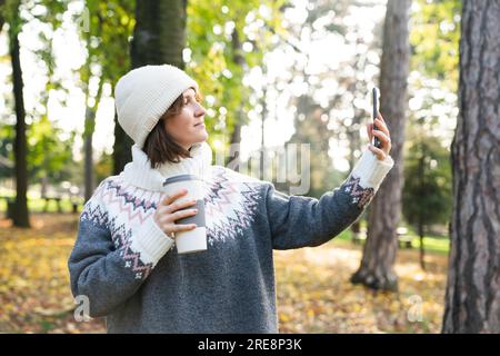 A woman is holding a phone and a cup of hot drink. Woman taking selfie on smartphone Stock Photo