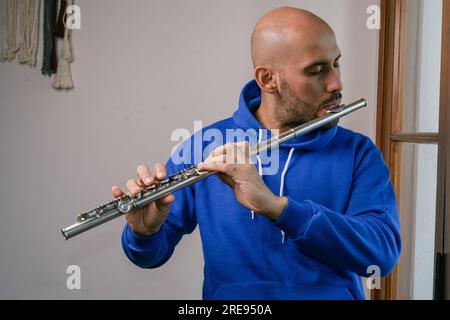 portrait of young latino man in blue, with beard and bald head standing at home playing flute with inspiration Stock Photo