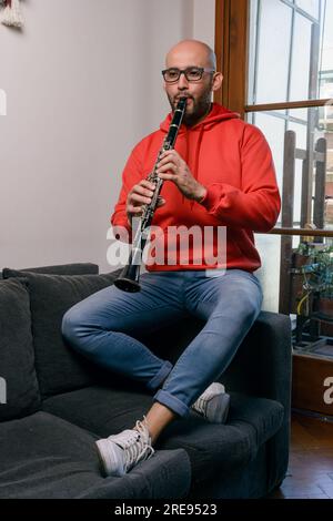 young latino man in red and blue, at home sitting on sofa in living room, playing clarinet, vertical image. Stock Photo