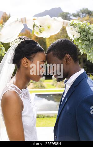 Happy african american bride and groom touching heads and smiling under wedding arch in sunny garden Stock Photo