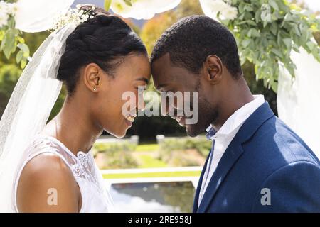 Happy african american bride and groom touching heads and smiling under wedding arch in sunny garden Stock Photo
