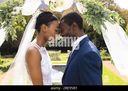 Happy african american bride and groom touching heads under wedding arch at ceremony in sunny garden Stock Photo