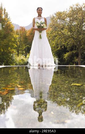 Happy african american bride holding bouquet, reflected in pond, in sunny garden, copy space Stock Photo