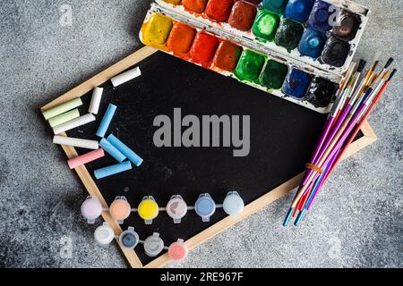 Top view of of used colorful aquarelle palette with bunch of paintbrushes placed on blackboard with chalks on gray background Stock Photo