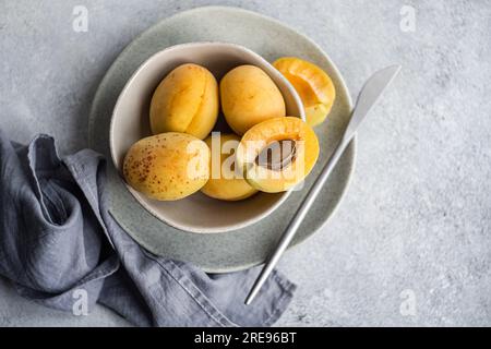 Top view of ceramic bowl with fresh ripe yellow apricots placed on blue cloth on concrete table with cut in half apricot Stock Photo