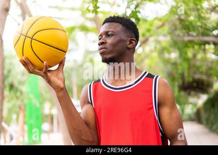 Portrait of confident young African American sportsman in activewear holding basketball while looking at it near blurred trees Stock Photo