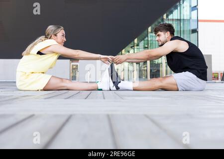 Side view of young couple looking at each other while sitting outside on paved floor and exercising in daylight with stretching of legs holding hands Stock Photo
