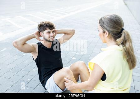 High angle of happy young couple in casual clothes smiling and looking at each other while doing sit up exercise on street pavement outdoors Stock Photo