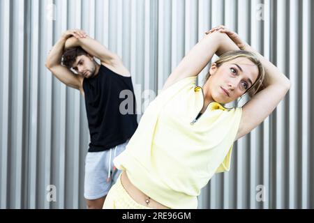 Serious young couple in casual clothes doing exercise while standing and swinging sideways on street by wall in daylight Stock Photo