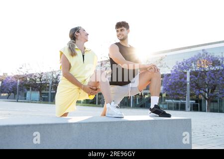 Happy young couple in casual sport clothes standing while stretching leg on concrete bench and looking at each other while exercising on city street Stock Photo