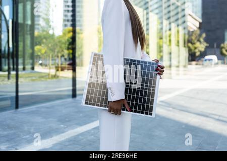 Side view of crop anonymous businesswoman in white blazer standing holding on street with portable solar panel on sunny day Stock Photo