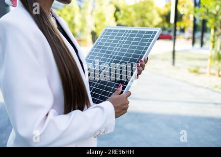 Side view of crop anonymous businesswoman in white blazer standing on street with portable solar panel on sunny day Stock Photo