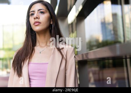 From below pensive young Middle Eastern businesswoman in formal clothes standing near urban glass building and looking away Stock Photo