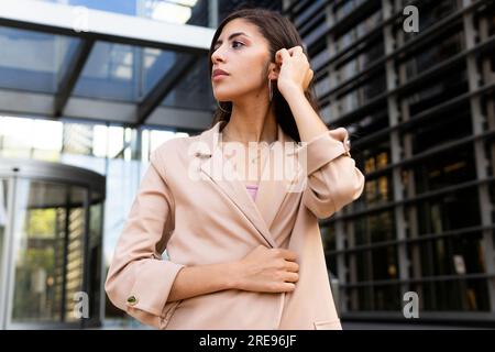 From below pensive young Middle Eastern businesswoman in formal clothes standing near urban glass building and looking away Stock Photo