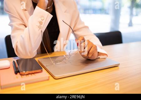 Crop anonymous businesswoman in white blazer sitting at wooden table near smartphone and laptop while holding eyeglasses in office Stock Photo
